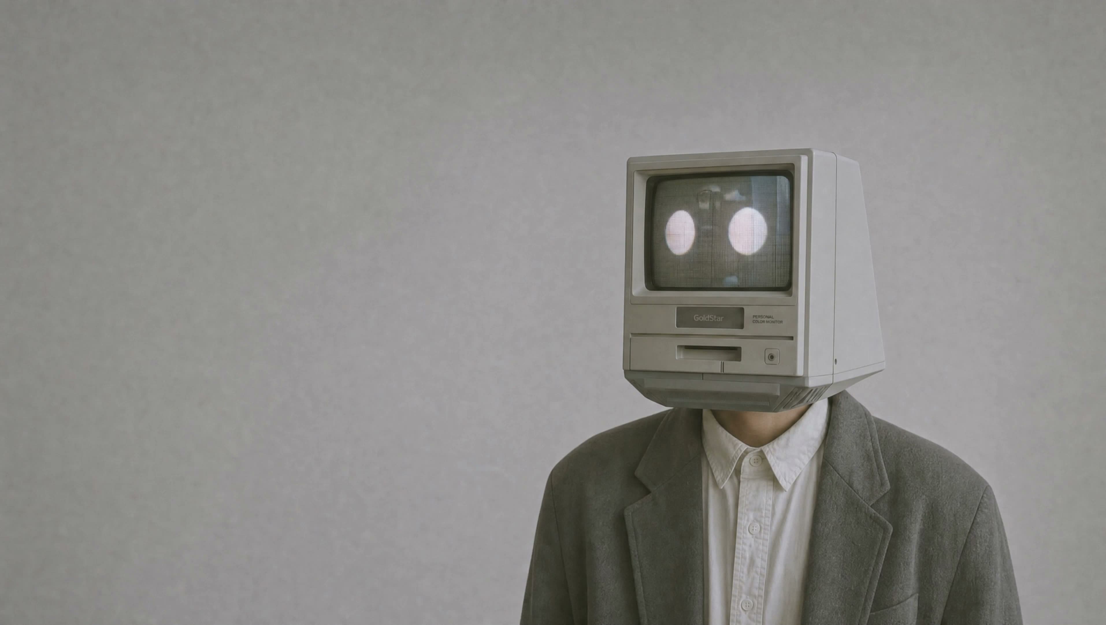 A figure in a gray blazer with a vintage computer monitor for a head, turning to follow your cursor